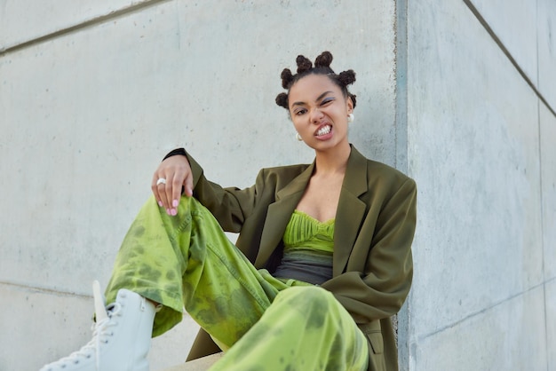 Cheeky fashionable brunette woman wears stylish green outfit white boots has vivid makeup sits outdoors near grey concrete wall clenches teeth feels self confident People and style concept