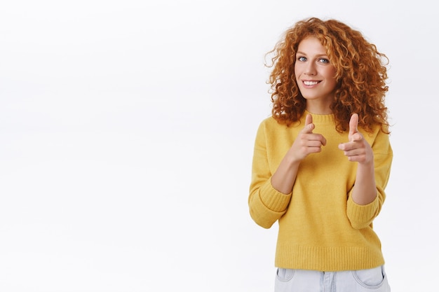 Free photo cheeky, friendly good-looking redhead curly woman in yellow sweater, pointing finger pistols at camera and smiling, picking you, playfully greeting or congratulating person, white wall