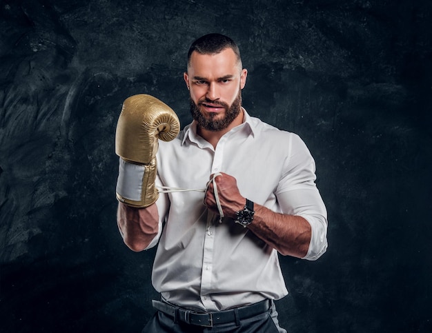 Cheeky handsome man in white shirt and with golden boxing glove is standing at dark studio.