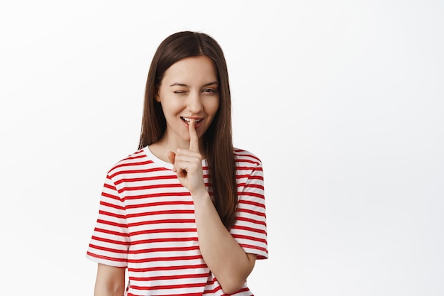 Cheeky young woman wink and hush, make shh gesture with finger near lips while smiling, has surprise, hinting at smth, standing in red t-shirt against white background