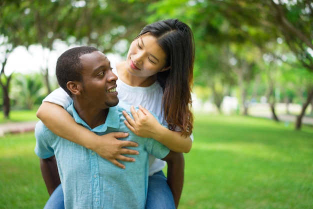 Cheerful African American man carrying Asian woman piggyback. 