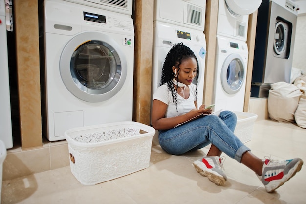 Cheerful african american woman sitting with earphones and read magazine near washing machine in the selfservice laundry