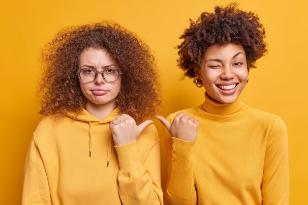 Cheerful Afro American woman and her sad curly haired sister point thumbs at each other express different emotions dressed casually isolated over yellow wall. Its she. Two women indoor