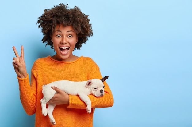 Cheerful Afro American woman makes peace gesture, enjoys good day, carries sleeping puppy