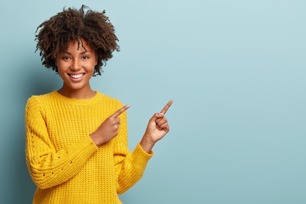 Cheerful Afro woman points away on copy space, gives way or direction, wears yellow warm sweater, has pleasant smile, feels optimistic, isolated over blue background