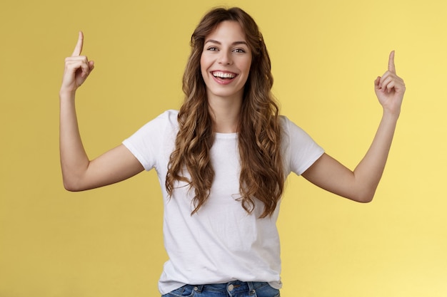 Cheerful amused happy lively young girl long curly haircut raise hands pointing up smiling toothy happily camera introduce excellent variant suggest you click site link advertising yellow background