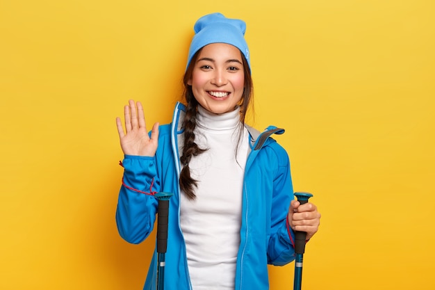 Cheerful Asain female traveler holds hiking equipment, waves palm, greets friend in mountains, being active hiker, smiles pleasantly, isolated over yellow wall