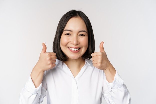 Cheerful asian woman face showing thumbs up and smiling pleased by something recommending smth good standing over white background