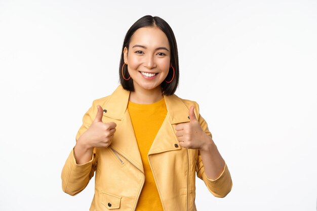 Cheerful asian woman smiling pleased showing thumbs up in approval standing in casual clothes over white background