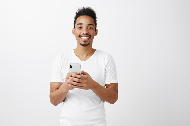 Cheerful attractive african american guy in white t-shirt holding mobile phone, smiling