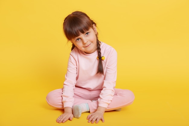 Cheerful attractive little girl sitting on floor with crossed legs, touching floor with palms, looking at camera, posing isolated over yellow background, dresses pale pink attire.