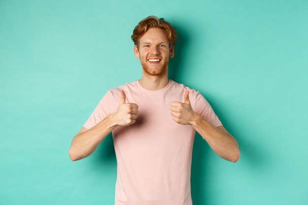 Free Photo cheerful bearded man with red hair showing thumbs-up, like and approve something, praising promo, standing against turquoise background