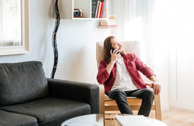 Free photo cheerful bearded young man sitting and talking on cell phone in armchair at home