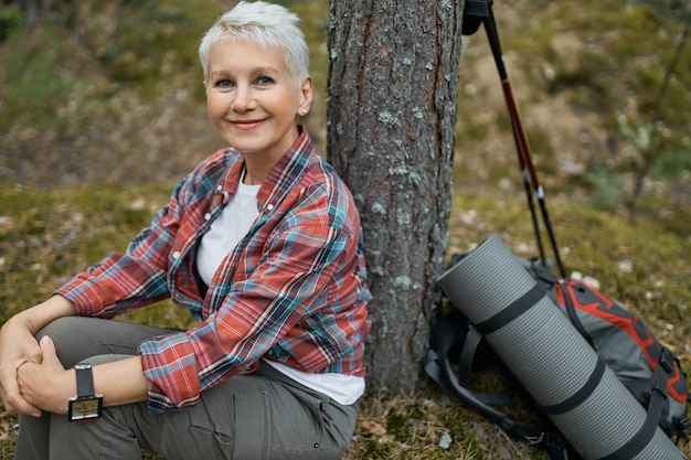 Cheerful beautiful female pensioner sitting under tree with backpack and sleeping mat, relaxing during her journey in wild nature. Attractive mature female having rest while hiking in forest