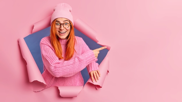 Free Photo cheerful beautiful millennial woman with natural red hair has beaming smile excited face expression pointing at copy space