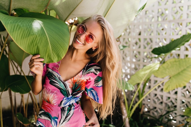 Free Photo cheerful blonde girl posing with sincere smile near big green leaf.