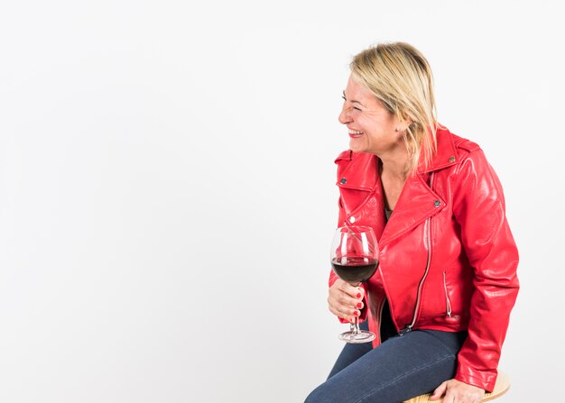 Cheerful blonde mature woman sitting on stool holding red wine glass in hand