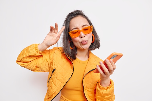 Cheerful brunette Asian woman dance to favorite song moves to rhythm of music holds modern smartphone wears trendy orange sunglasses stylish jacket expresses happiness and joy models indoor.