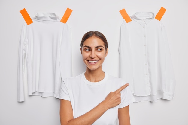 Free Photo cheerful brunette european woman points away with index finger demonstrates something advertises clothes dressed in casual t shirt poses against white background two shirts plastered behind