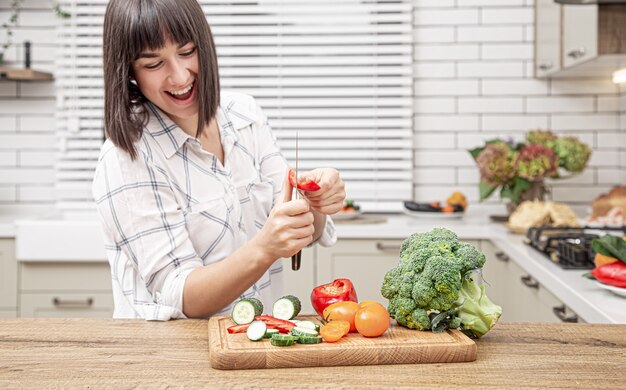 Cheerful brunette girl cuts vegetables on salad on the background of modern kitchen interior.