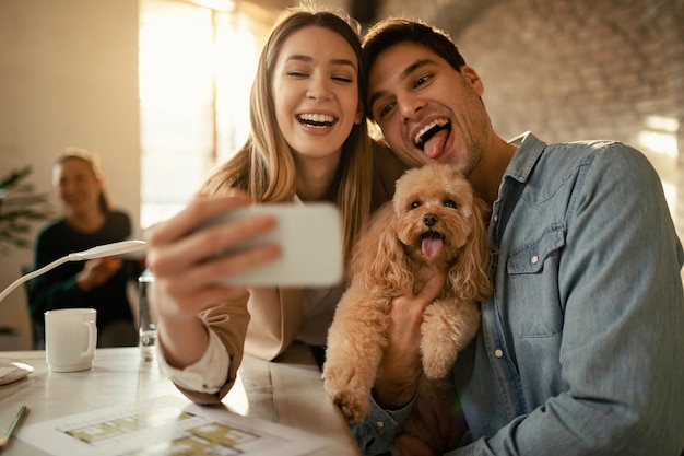 Free photo cheerful business couple having fun with their dog and taking selfie with mobile phone in the office