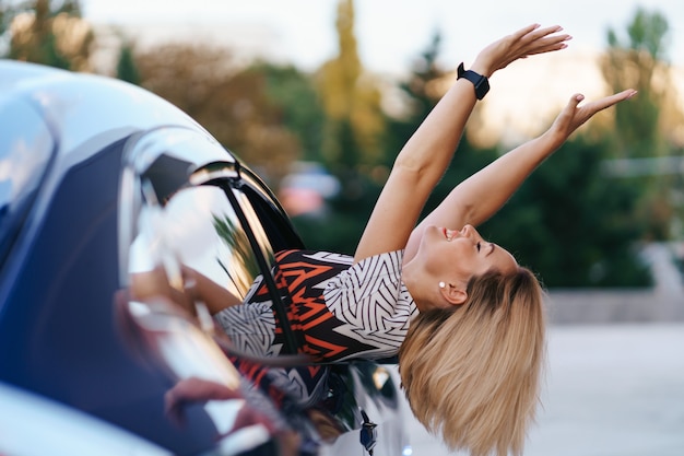 Free photo cheerful caucasian woman drives through the picturesque sunny city and waves her arms while stretching out of the car window on a beautiful day