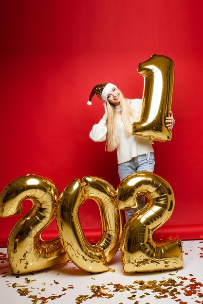 Cheerful caucasian young woman in white sweater, blue jeans and red christmas hat smiles, picture isolated on red background