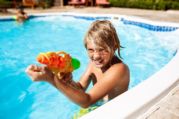 Cheerful children playing waterguns, rejoicing, jumping, swimming in pool.