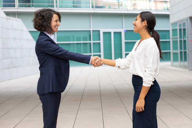 Cheerful colleagues shaking hands near office building. Young women wearing formal suits meeting outdoor. Business handshake concept