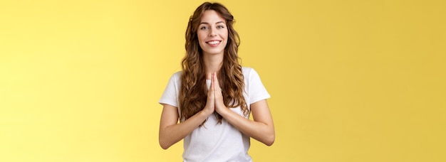 Free photo cheerful confident outgoing young polite girl say namaste greeting asian guests smiling gladly thanking for visit hold hands pray supplication gesture asking favour begging stand yellow background