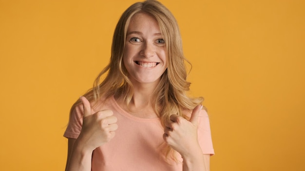 Free Photo cheerful cool blond girl keeping thumbs up joyfully looking in camera isolated on yellow background like expression