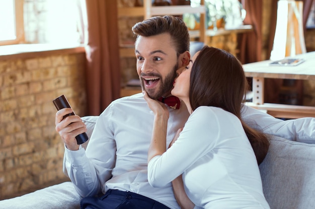 Cheerful couple watching television. Beautiful woman kissing man on couch in livingroom.