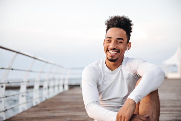 Cheerful curly dark-skinned man in white long-sleeved t-shirt smiles sincerely and rests near sea