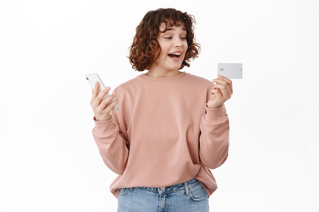 Cheerful curly girl, looks excited at credit card, holds smartphone in hand, makes online order, shopping in internet store, standing happy against white background