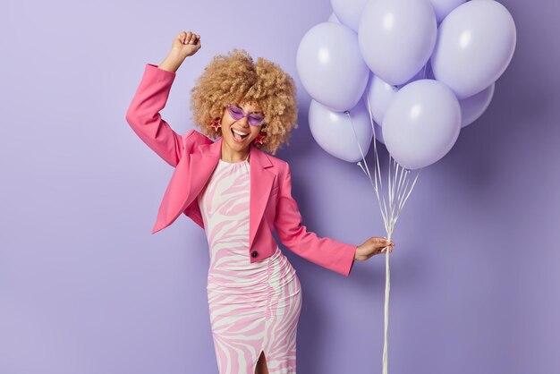 Cheerful curly haired beautiful woman dances with rhythm of music dressed in festive clothes enjoys special occasion holds bunch of balloons isolated over purple background Party time concept