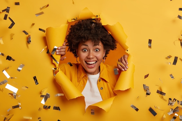 Free Photo cheerful curly haired lady with toothy smile, wears fashionable clothes and silver earrings, looks through paper hole with bright sparkles