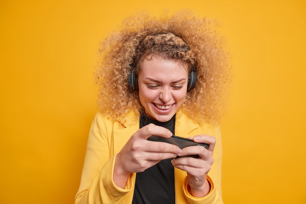 Free Photo cheerful curly haired woman addicted to video games plays on mobile phone uses wireless headphones concentrated at display enjoys gaming dressed formally isolated over vivid yellow wall