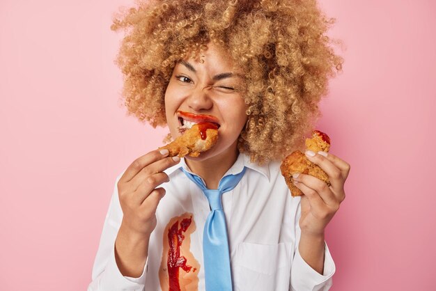 Cheerful curly haired woman eats delicious meal fast food smeared with ketchup keeps to unhealthy nutrition has binge eating habit wears white shirt and blue tie isolated over pink background