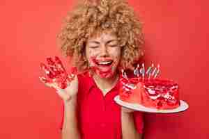 Free photo cheerful curly haired woman foolishes around dirty with cream eats tasty sweet cake with hands keeps palm raised exclaims from joy wears dress isolated over vivid red background dessert eating