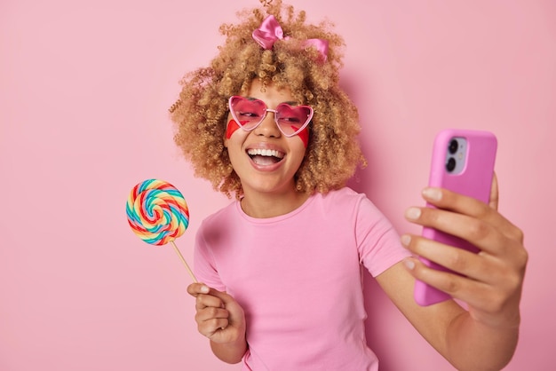 Free photo cheerful curly haired woman holds big rainbow caramel candy on stick laughs gladfully takes selfie via smartphone foolishes around wears sunglasses and casual t shirt isolated on pink background