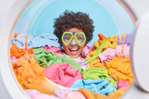 Free photo cheerful curly haired young busy woman wears snorkeling mask for diving has lot of laundry to make does housework drowned in multicolored clothes against blue wall