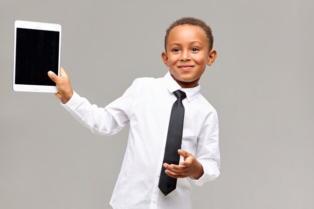 Cheerful cute Afro American schoolboy in shirt and tie smiling happily using electronic gadget to play games or watch cartoons, holding digital tablet with blank display with copyspace for your text