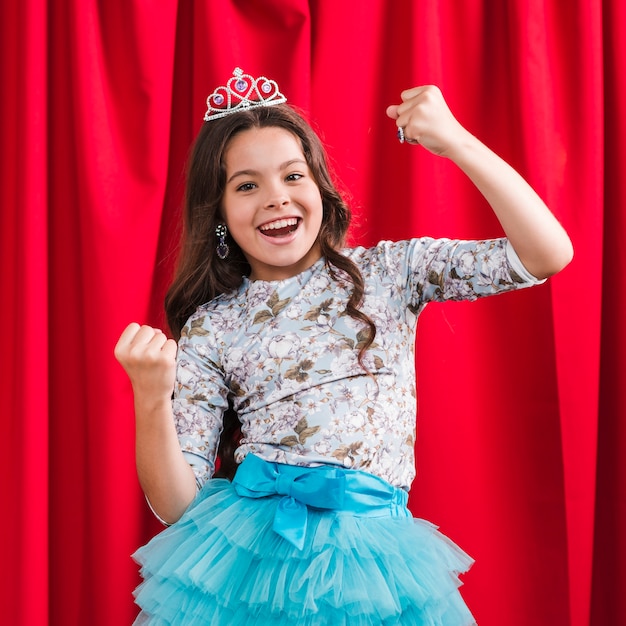 Cheerful cute girl making winner gesture standing in front of red curtain