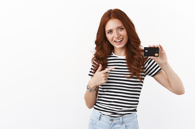 Free photo cheerful cute redhead girl opened deposit gather money for summer holiday, pointing at black credit card and smiling joyfully, paying online, using cashback to pay for coffee