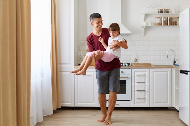 Cheerful dad carrying his dark haired daughter, playing with happy little preschool child indoors against kitchen set, playful small kid girl having fun with smiling father at home.