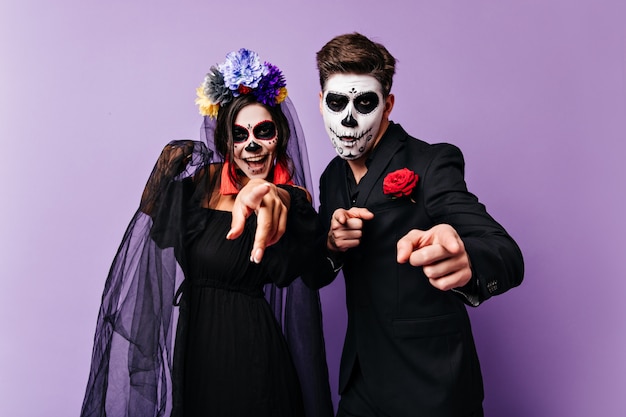 Cheerful dark-haired boy and his girlfriend with faces painted for Halloween are smiling and showing fingers to camera.