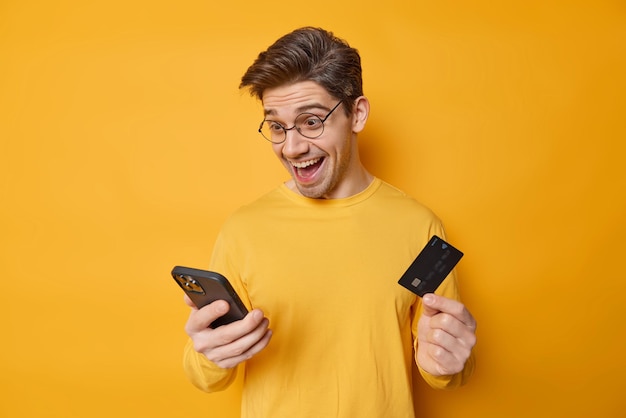 Free Photo cheerful dark haired man holds smartphone and credit card involved in online shopping purchases goods or purchases in internet store wears spectacles casual jumper isolated over yellow wall.