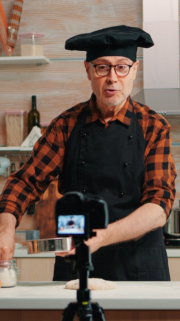 Free photo cheerful elderly baker man filming cooking vlog in home kitchen. retired blogger chef influencer using internet technology communicating, shooting blogging on social media with digital equipment