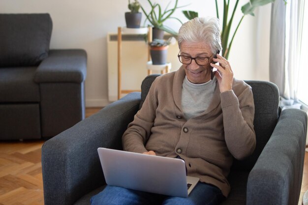 Cheerful elderly freelancer working remotely. Smiling gray-haired man in cardigan using laptop while calling by phone. Online business concept