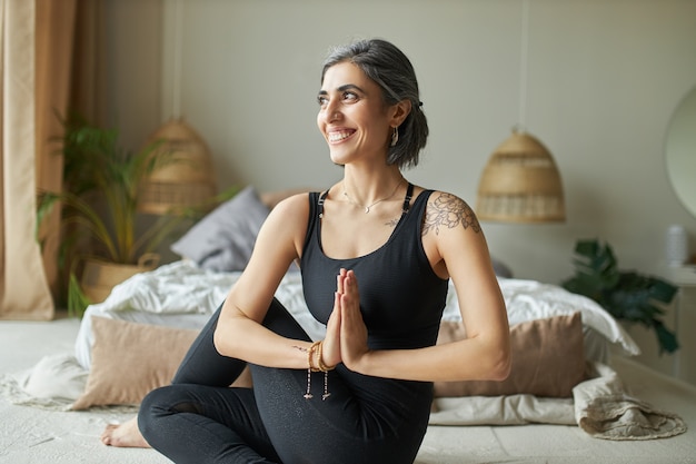 Free Photo cheerful energetic young female sitting in spinal twist on floor at home, doing ardha matsyendrasana during yoga practice